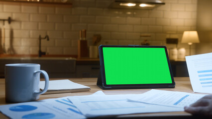 Person working on tablet computer with green mock up screen on table in the kitchen. Co-workers working with papers, remote work, home office concept	