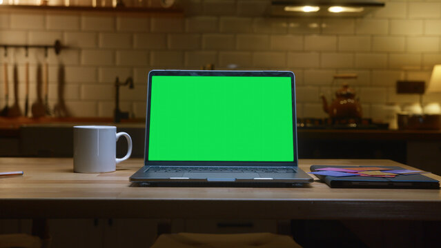 A laptop with a green screen on a kitchen table in kitchen with a coffee mug and notebook.