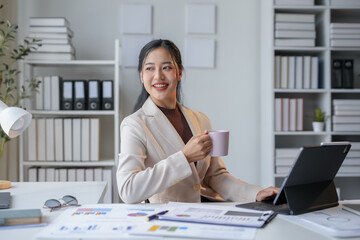 Young manager is smiling while working on a tablet and drinking coffee at her desk in a luminous...