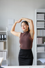 Young asian businesswoman is stretching her arms at office desk, enjoying break at work and feeling relaxed