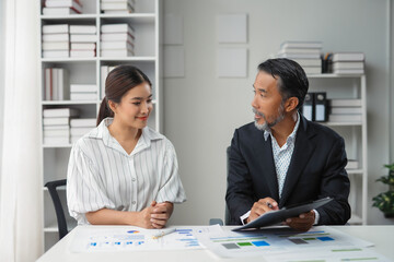 Manager holding a file discussing charts with a young intern while sitting at an office desk