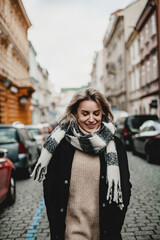 A woman wearing a black and white scarf is walking down a city street. The street is lined with parked cars, including a red car. The woman is smiling and enjoying her walk