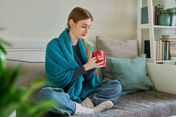 Young woman warming under blanket with mug of hot drink sitting on couch at home
