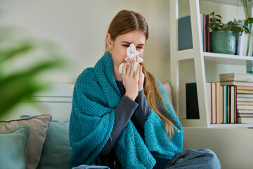 Young sick woman sitting under warm blanket sneezing coughing in handkerchief