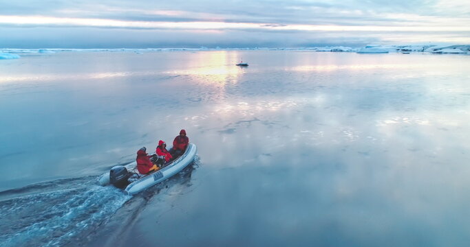 Group of scientists ride motorboat in Antarctica. Zodiac boat glides on ocean surface in sunset light. Antarctic travel and expedition. Discover beauty of South Pole. Aerial drone winter landscape