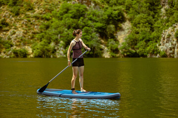 Woman paddles on lake or river standing on SUP wearing a two piece bathing costume with shorts; slim athletic brunette female on a stand up paddleboard in summer sunshine with trees in background.