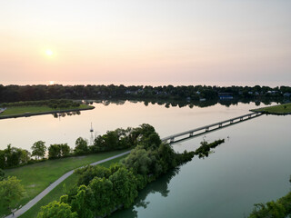 Aerial view of a pedestrian bridge connecting Rennie Park and Royal Henley Park across Martindale Pond in St. Catharines, Ontario, Canada during a hazy orange summer sunset, July, 2024.