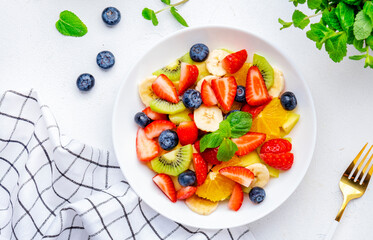 Summer vegan fruity salad with strawberries, blueberries, banana, kiwi, orange and mint on white background, top view