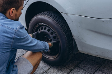 Young man in unscrewing lug nuts on car wheel in process of new tire replacement,using wrench while...