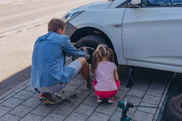 Cute little girl helps her father to change wheel on their family car on warm day in the yard