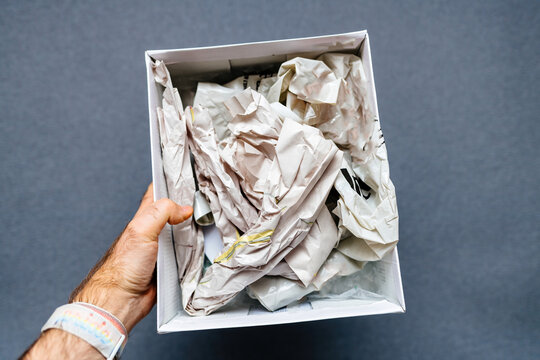 A male hand holding a paper box filled with newspaper against a gray background, in the process of unboxing a fragile object, showcasing careful handling and protection
