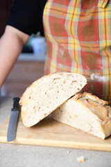 Person with an apron standing next to a cut loaf of bread on a cutting board.