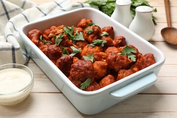 Baked cauliflower buffalo wings with parsley in baking dish and sauce on wooden table, closeup