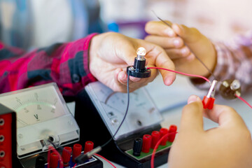 Children are studying electrical circuit systems by experimenting with electrical connections using a small electric meter and a small flashlight bulb in physics science class, soft focus.