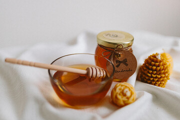 Organic honey in a glass jar and a glass small bowl and burning candles made of natural handmade wax on a white background. Beekeeping products. Small business. Farm.