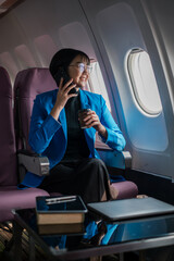 Professional woman in blue blazer using smartphone and drinking coffee on airplane. Business travel concept.