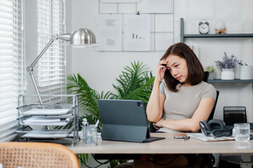 Young woman in home office, appearing stressed while working on a laptop. Modern workspace with plants, shelves, and office supplies.