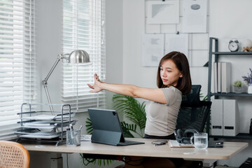 A young woman stretches at her desk in a modern home office, surrounded by office supplies and a...