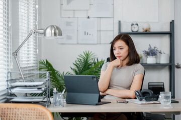 A young woman working on a tablet in a modern home office setting, surrounded by plants and shelves, looking focused and thoughtful.