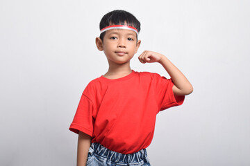 Confident Asian boy in red t-shirt flexing strength by raising his arms and muscles, isolated on white background. Indonesian Independence Day concept.