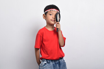 Smart Asian boy holding magnifying glass to search information, isolated on white background. Indonesian Independence Day concept.