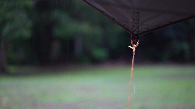 camping on rainy season with closeup rain water drips droplets to tip tarp and flow motion on fly sheet tent with carabiner paracord rope for holiday vacation picnic on green nature forest garden