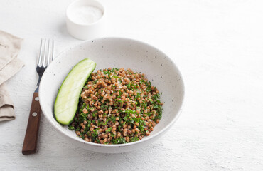 White bowl with boiled buckwheat and kale on a gray background, served with fresh cucumber. Healthy homemade vegan food