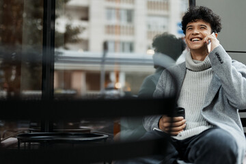 Stylish man sitting in a chair by the window, talking on his cellphone with a coffee cup in his hand