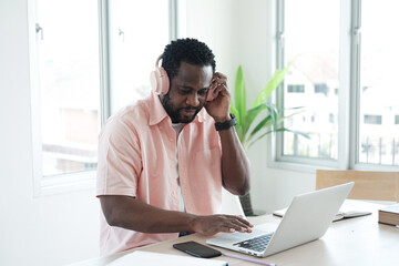 Fototapeta premium Smiling African American businessman is listen to online music and meeting headphone. man searching internet on laptop in modern office. Business and technology concept