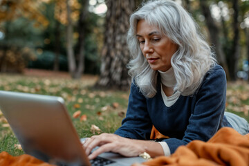 Middle-aged woman with silver hair working on a laptop in a park during autumn