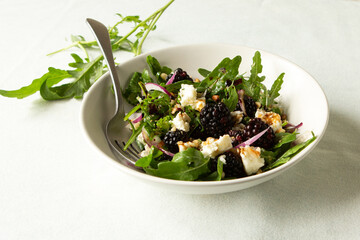bowl of salad with arugula, blackberries and feta cheese on white table