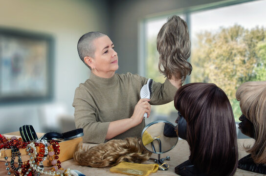 Senior woman caring for wigs in a bright room, holding one at eye level while others are displayed on mannequin heads.