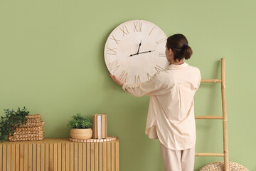 Young woman hanging clock on green wall near chest of drawers in room