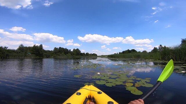 Kayaking along river through growing white lily nymph on sunny summer day. Person floating in boat on river with surface of water reflecting white clouds on calm day. Tourism. Active lifestyle. POV