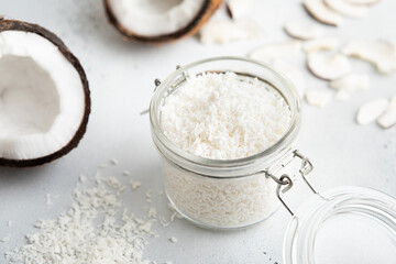 Shredded sweet coconut flakes in glass jar with ripe coconuts on light table.Macro
