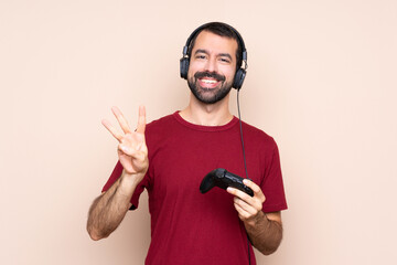 Man playing with a video game controller over isolated wall happy and counting three with fingers