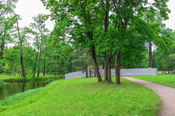 A winding path leads through a verdant forest, flanked by a tranquil river. Sunlight filters through the dense canopy of trees, casting dappled shadows on the grassy banks.