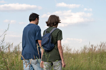 Man and teenager boy walking in tall grass in meadow, father and son hiking together outdoor, back view, blue cloudy sky background