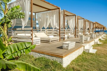 Sunny beach club with a row of luxury gazebos featuring white curtains, wooden decks, and lush...