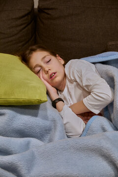 Vertical photo of 10-year-old girl sleeping on the sofa with a green cushion as a pillow and covered with a blue blanket