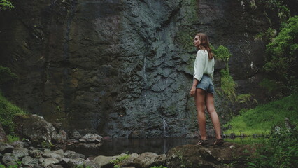 Naklejka premium A woman stands admiring a waterfall in French Polynesia, her back to the camera, as she gazes at the cascading water and lush greenery.