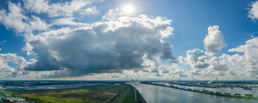 Massive cloud formations are present above the entrance of the Port of Rotterdam with greenhouses of the Westland region on the left, wind turbines in the middle, and oil refineries on the right.