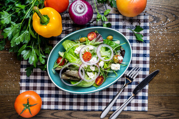 Toss The Green salad with spinach, cheese, olives, cherry tomato, tomato, onion, capsicum, coriander, knife and fork served in dish isolated on napkin top view of italian healthy food on wooden table