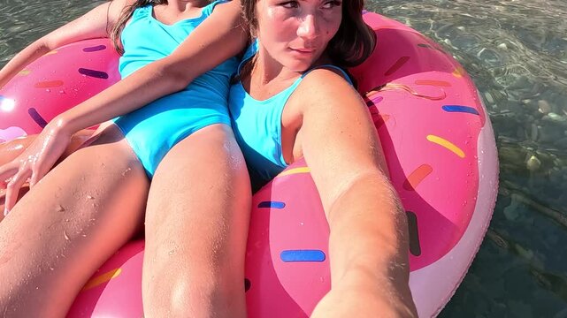 Two women in blue swimsuits float on a pink donut-shaped inflatable ring in the water. One woman is taking a selfie. Summer holiday vacation and travel concept