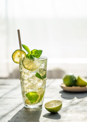 Mojito cocktail in a tall glass with lime, mint and ice on a table with morning light and shadow.