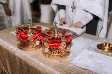 Two beautiful Christian crowns made of red fabric, with a gold frame, a pattern, and a cross lie on the table at a wedding ceremony, a rite in the church. Photography, religion.