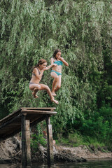 Two girlfriends, athletic girls, children jump from a wooden bridge, pier into the water on a lake outdoors in summer, holding hands together. Photography, portrait, friendship concept, sport.