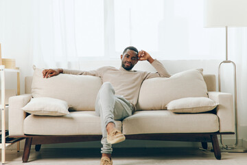 Obraz premium Man Sitting on Sofa at Home, Lost in Sad Thoughts, Finding Comfort and Relaxation A young African American guy in a modern living room, smiling and watching something online, holding a smartphone His