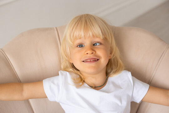 Little happy smiling boy at home with strabismus eyes, squinting eyes, cross-eyed. Child boy wearing white t-shirt on a beige chair near white wall