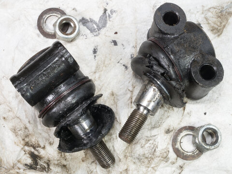 Closeup overhead shot of two worn and oily suspension ball joints, lying on dirty paper, which have been removed from a vehicle for replacement.